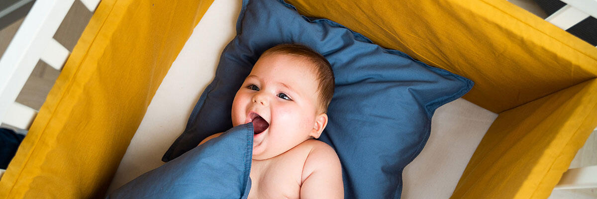 Baby som ligger i barneseng med sennepsgul sengekant og blått sengetøy. Bilde