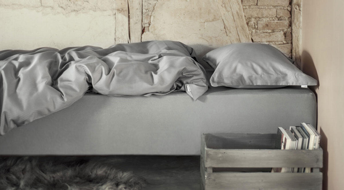 Seng ferdig oppredd med laken, sengetøy og putevar i lys grå farge. Bilde